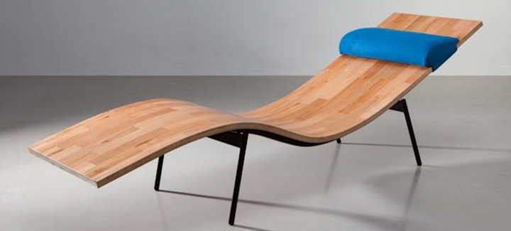 Moderne Relaxliege aus geformtem Holz mit schwarzem Gestell und blauem Kopfpolster in Galerieumgebung.